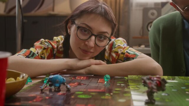 Low angle portrait shot of calm and smiling young woman in glasses resting her head on her hands while participating in RPG tabletop game with friends