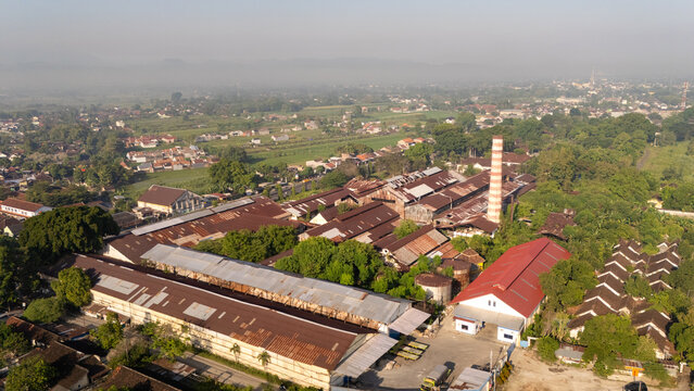 Aerial view of an old industrial factory complex with rusted metal roofs and a tall brick chimney, surrounded by lush green trees and rural landscape in Indonesia.