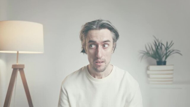 Young man in white sweater sitting indoors in minimal room, looking at camera with varied expressions, illuminated by soft daylight and warm lamp