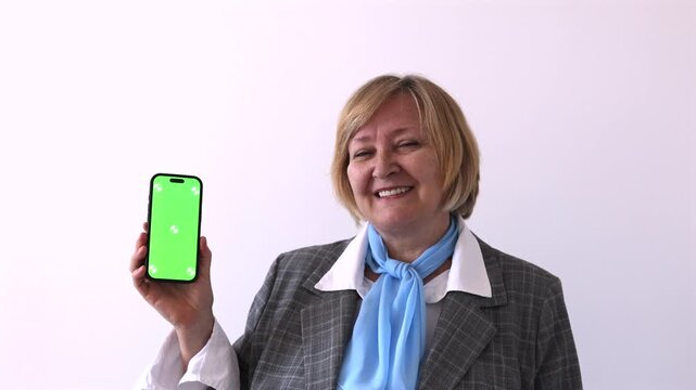 Senior Businesswoman Showing Green Screen Smartphone. Smiling senior businesswoman holding a smartphone with green screen for app, website or mobile interface mockup against a plain light background.
