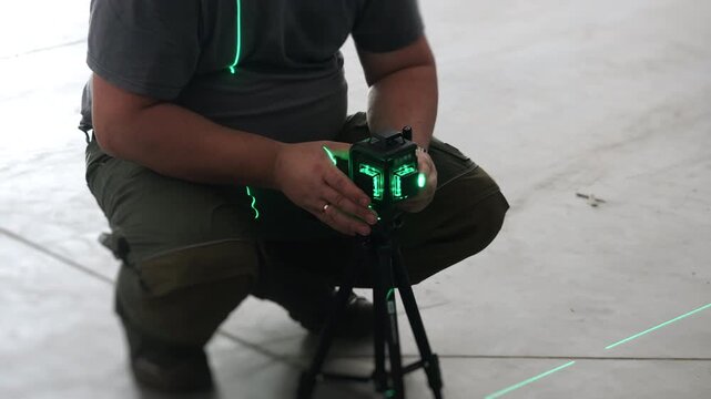 Worker adjusting a green laser level mounted on a tripod inside an industrial warehouse before installation, construction and precision alignment work.