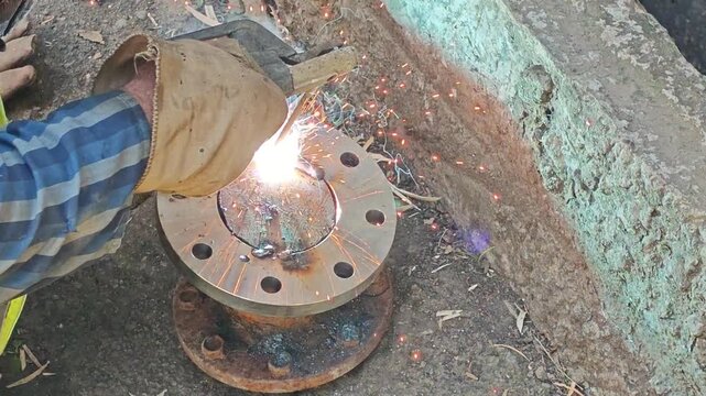 Close-up of a welder welding a steel pipe flange with an electrode, creating bright sparks and smoke.