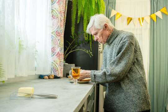 An elderly man with silver hair stands at kitchen counter, having prepared tea and butter sandwiches. The image conveys quiet satisfaction of self-prepared meals of senior. Grandpa before eating.