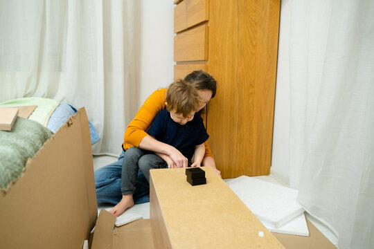 A woman assembles a cabinet in the room while holding her child on her lap. Balancing parenting and household duties, showcasing independent motherhood and DIY skills.