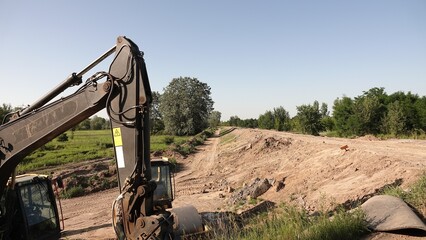 Pionowe ujęcie z bliska przedstawiające potężne ramię i łyżkę koparki na tle kołowej ładowarki i bezchmurnego nieba na placu budowy. © tomo81