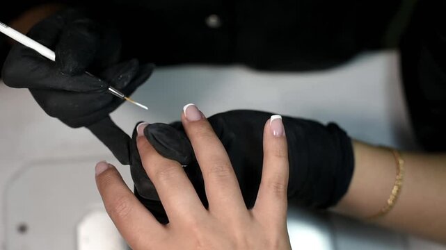 A professional nail technician wearing black gloves uses a fine detail brush to paint white tips for a classic French manicure. The close-up view highlights the meticulous process.