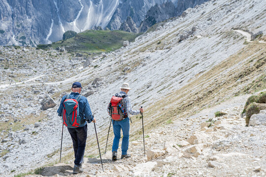 Two senior male hikers with backpacks and trekking poles walking down a steep gravel mountain trail, active aging and high-altitude alpine exploration concept