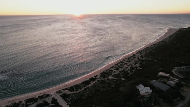 Waves roll onto the shore as the sun sets on the horizon, illuminating the water and the landscape around the beach.