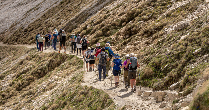 Group of tourists with backpacks and trekking poles walking along a busy mountain trail, active summer group travel and high-altitude alpine exploration concept