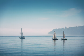 Fototapeta premium Sailboats With White Sails and With Crew Aboard Sailing on a Calm Foggy Sea Under a Pale Blue Sky Near the Horizon