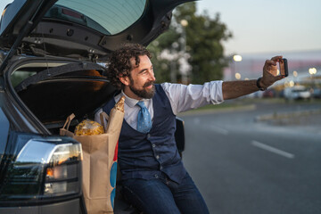 Naklejka premium Man taking selfie sitting in car trunk with groceries