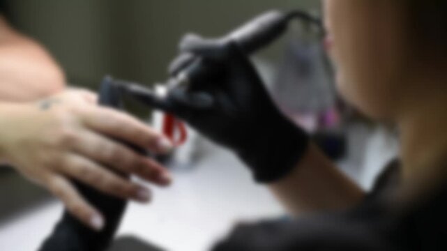 A professional manicurist uses an hardware electric nail drill tool to file a customer's fingernails. The soft-focus blurred shot captures the motion in a beauty salon background.
