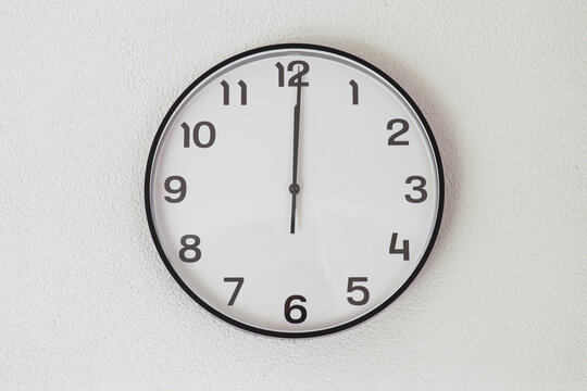 Twelve o'clock on black and white clock on white wall. Telling time on analogue clock with 12 numbers. It's 12 o'clock in the afternoon