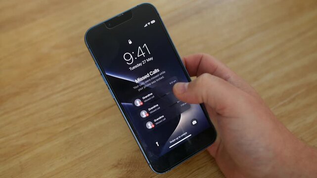 Phone showing missed calls from Grandma rests on a wooden table as a hand taps the lock screen. Close-up mobile, cell, smartphone view highlights granny, grandmother and family contact alerts