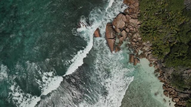 Experience the stunning beauty of Anse Cocos, La Digue island from above. Turquoise ocean waves crash along the rocky shore, revealing clear water and lush coastal vegetation in bright daylight.