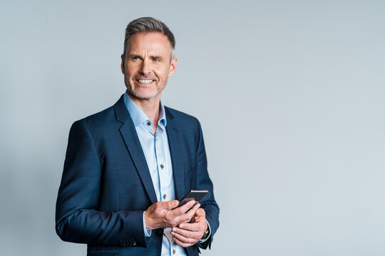 Smiling male entrepreneur holding smart phone while standing by gray background
