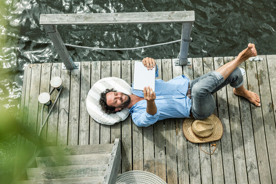 Man lying on platform at the waterside holding digital tablet