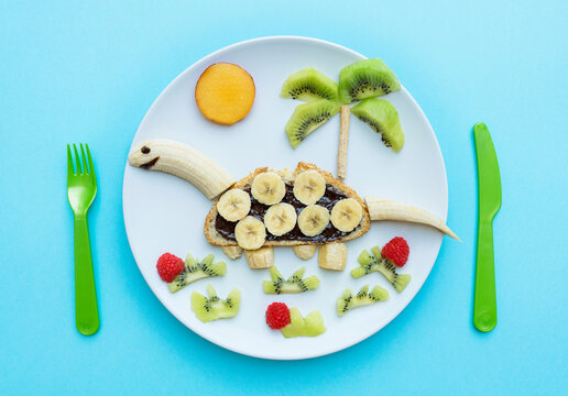 Breakfast with dinosaur shape made of banana, kiwi, bread, chocolate spread and berries on white plate on blue background