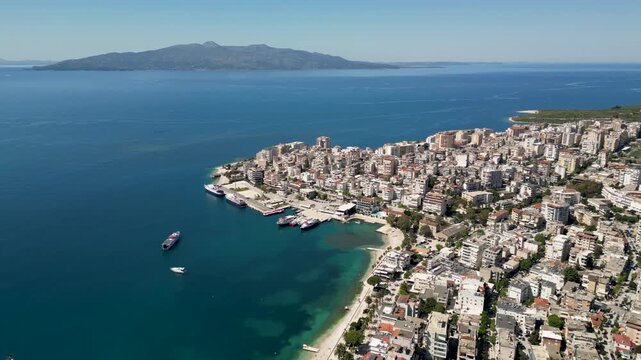 Drone footage capturing Saranda's harbor, waterfront promenade, urban coastline, and the Ionian Sea. Wide aerial panorama of the coastal city with Corfu Island visible across the water.