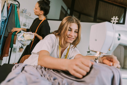 Smiling teenage fashion designer sewing cloths with mature woman in background working at workshop