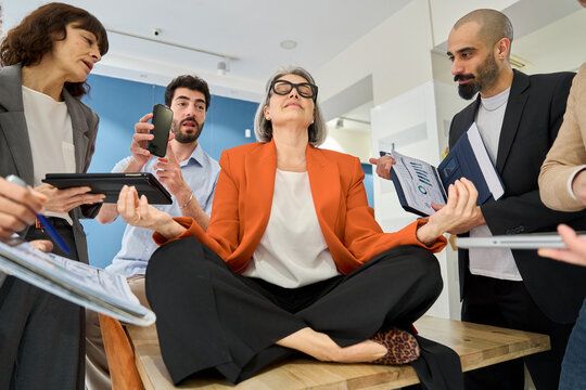 Businesswoman meditating on office desk among stressed coworkers