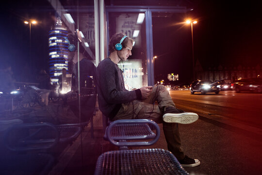 Germany, Munich, man with headphones sitting at bus stop using digital tablet at night