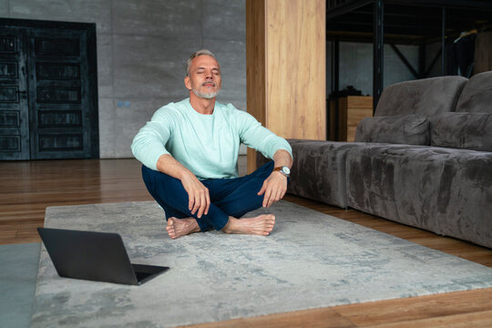 Man with eyes closed doing yoga by laptop on carpet at home
