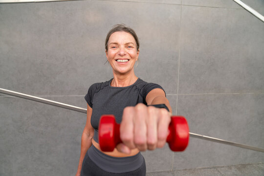 Happy woman showing red dumbbell in front of wall
