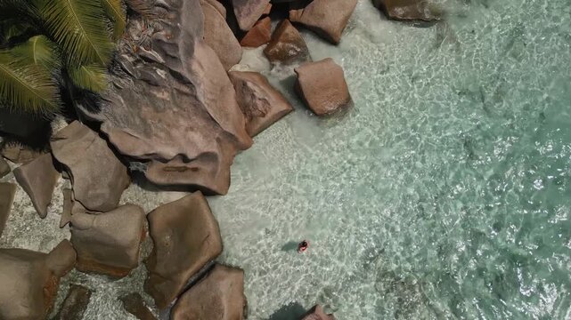 Woman in red swimsuit enjoys swimming in clear turquoise waters at Anse Cocos beach. Aerial view slowly rises, revealing granite rocks and lush vegetation on La Digue island.