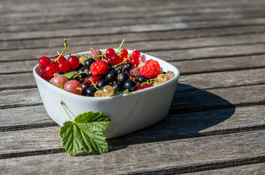 A white dish with a mix of fresh berries on wooden table, leaf close to dish
