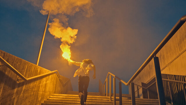 A young skateboarder strides up outdoor stairs clutching a flaming torch, exuding a carefree and rebellious vibe against a blue sky, embodying youthful exuberance.