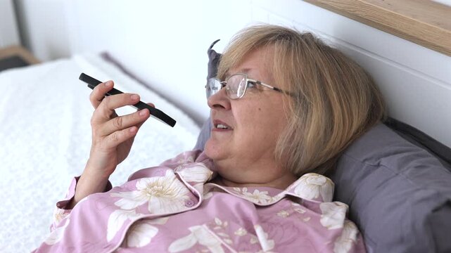 Senior Woman Recording Voice Message in Bed. Smiling senior woman in pajamas lying in bed and recording a voice message on smartphone at home.