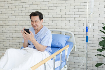Smiling Asian senior man patient using smartphone while sitting on hospital bed. Happy male...