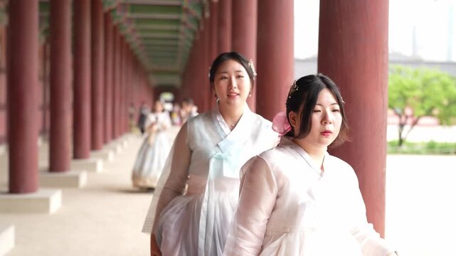 Two women in hanboks walk under pillars. One gestures toward distant scenery, smiling. The other looks forward, equally cheerful. Bright daylight highlights traditional architecture