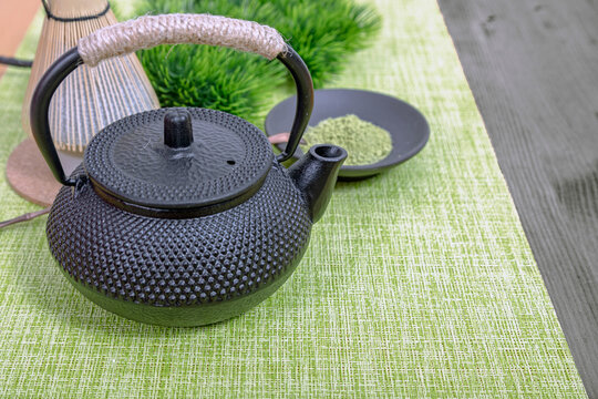 Cast iron teapot with matcha powder and bamboo whisk on green textured table runner. Traditional Japanese tea ceremony preparation with healthy organic green tea ingredients