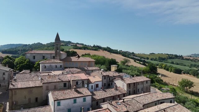 Villaggio rurale con edifici in pietra e tetti in coppi, dominato da un alto campanile, circondato da colline verdi e campi coltivati sotto cielo azzurro limpido