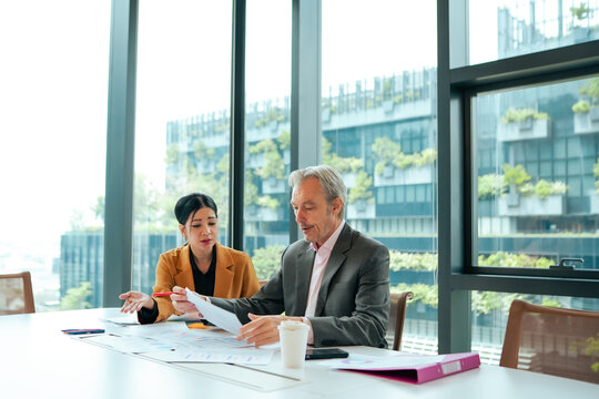 Senior executive reviews financial reports with young business professional during corporate meeting inside modern office. Business planning, document analysis, strategic decision making.