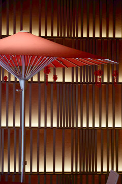 A red parasol with hanging Chinese decorations stands against a modern vertical wood slatted wall, lit by warm ambient lighting, creating a cozy, festive and contemporary interior scene..