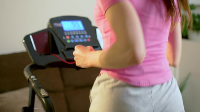 Back view of unrecognizable woman running on treadmill with digital console in home fitness space. Steady jogging motion during indoor cardio workout and daily endurance training routine. Healthy
