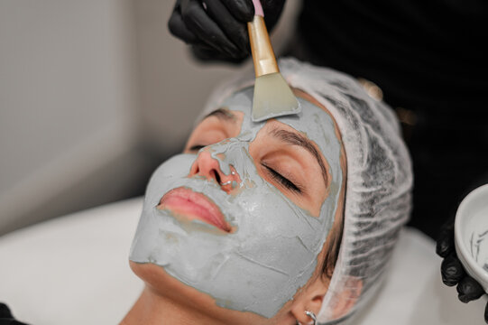 Beauty therapist applying a grey facial mask to a client lying on a treatment bed