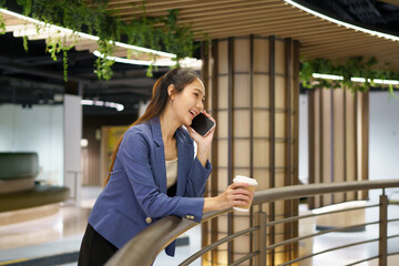 Fototapeta premium Young Asian Businesswoman Talking on Smartphone While Holding Coffee Cup in Modern Office Building