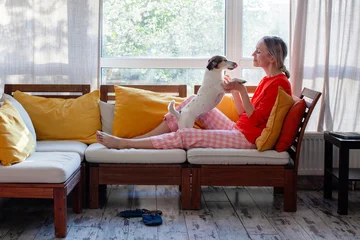 Woman relaxing on a wooden sofa and playing with her terrier dog at home. Leisure time with favorite pet in cozy living room. Domestic life and friendship between owner and animal. © Tatyana Gladskih