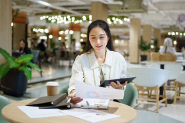 Obraz premium Young Asian Businesswoman Working with Tablet and Documents at Modern Office Desk in cafe with tablet