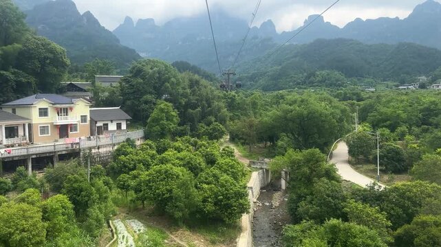 Aerial view of cable car riding over green mountain landscape. Gondola lift transport system in valley near village. Travel and excursion in nature, scenic mountain sightseeing location.
