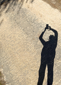 Silhouette of person standing with arms raised taking a photo selfie with a mobile cell phone shadow and concrete monotone background