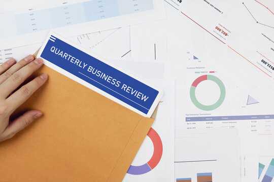Hand pulling a quarterly business review document out of a brown envelope on financial charts. Concept of performance evaluation, corporate data analysis, and strategic report.