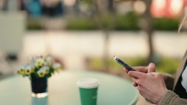 Close-up of a young woman's hands using a mobile phone while sitting at a table with coffee and flowers in an outdoor cafe, enjoying a relaxing break and communicating on social media