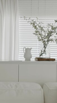 Calm and serene living room and kitchen room featuring a bright, minimalist all-white interior with a plant in a glass vase by the window, peaceful clean aesthetic modern home decor. vertical video