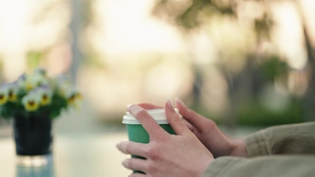 Woman with a beautiful manicure picking up a green paper coffee cup from a glass table at a sunlit outdoor cafe, with a vase of flowers in the blurred background creating a relaxing atmosphere