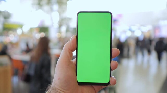 Hand holds phone with green screen. Background blurred, public space visible. Ideal for app mockups or digital content. People appear passive, not interacting with device. Perfect for advertising